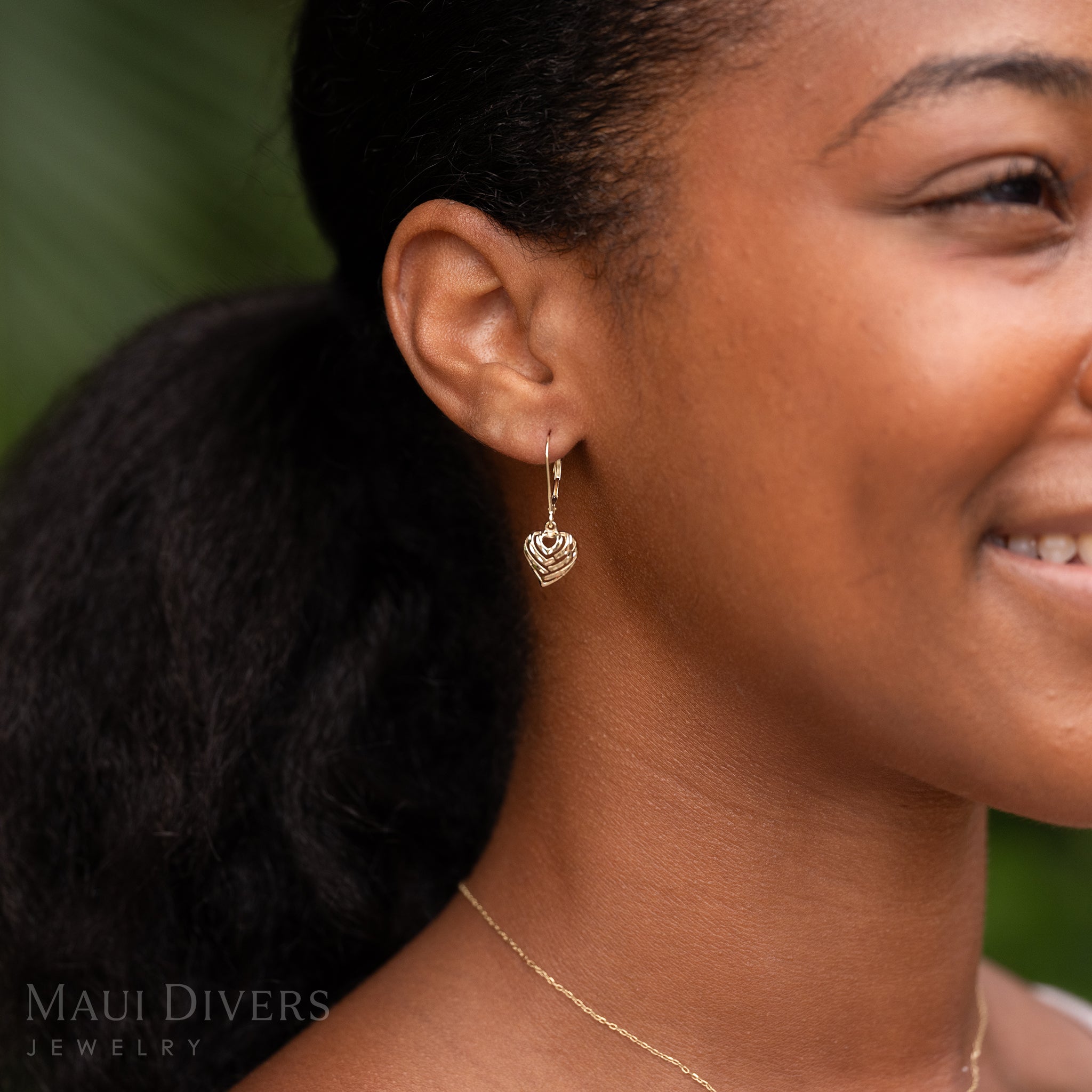Close-up of a smiling woman wearing an Aloha Heart Earring with Diamond-cut finish in 14k Yellow Gold against a blurred leafy green background