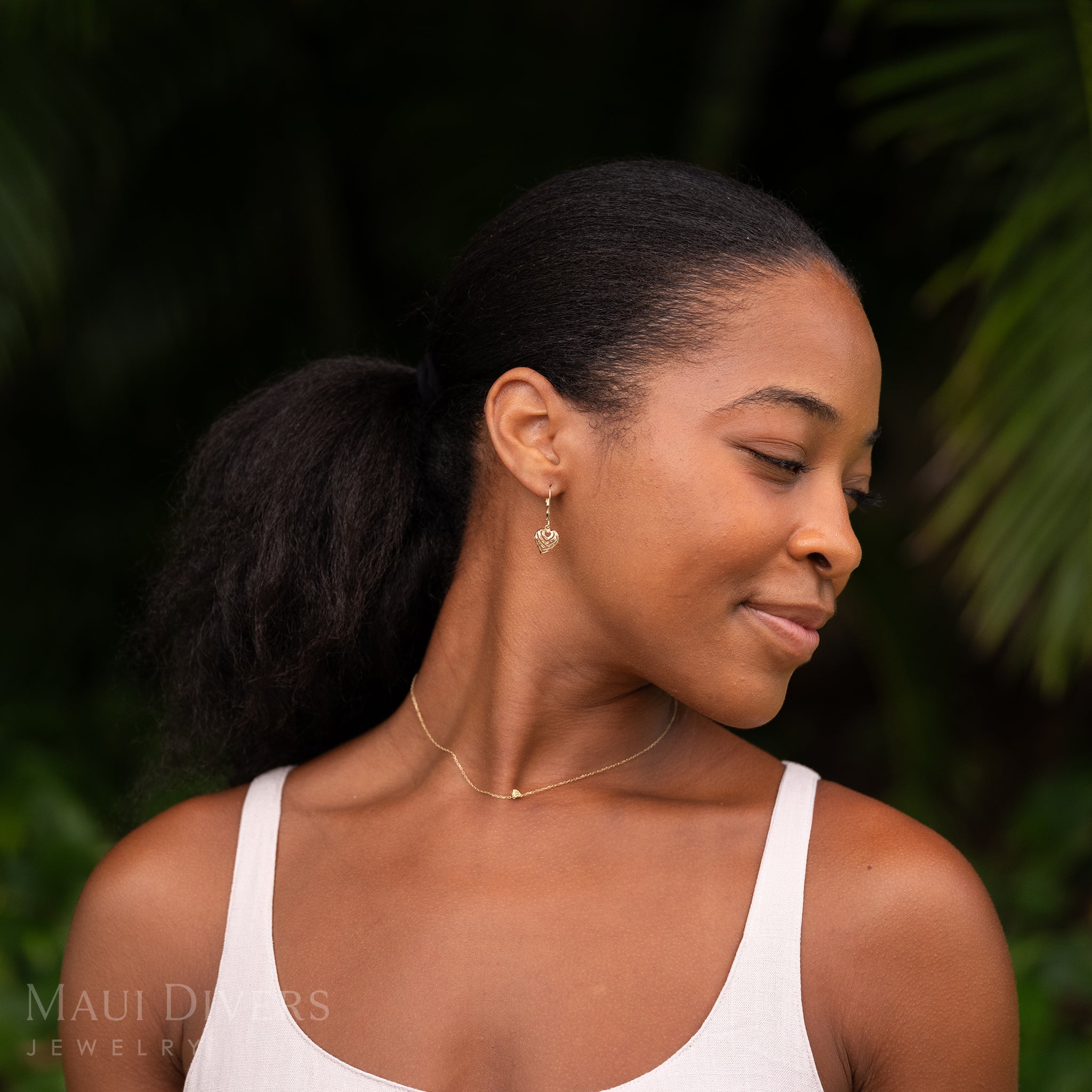 A smiling woman wearing an Aloha Heart Earring with Diamond-cut finish in 14k Yellow Gold against a blurred leafy green background