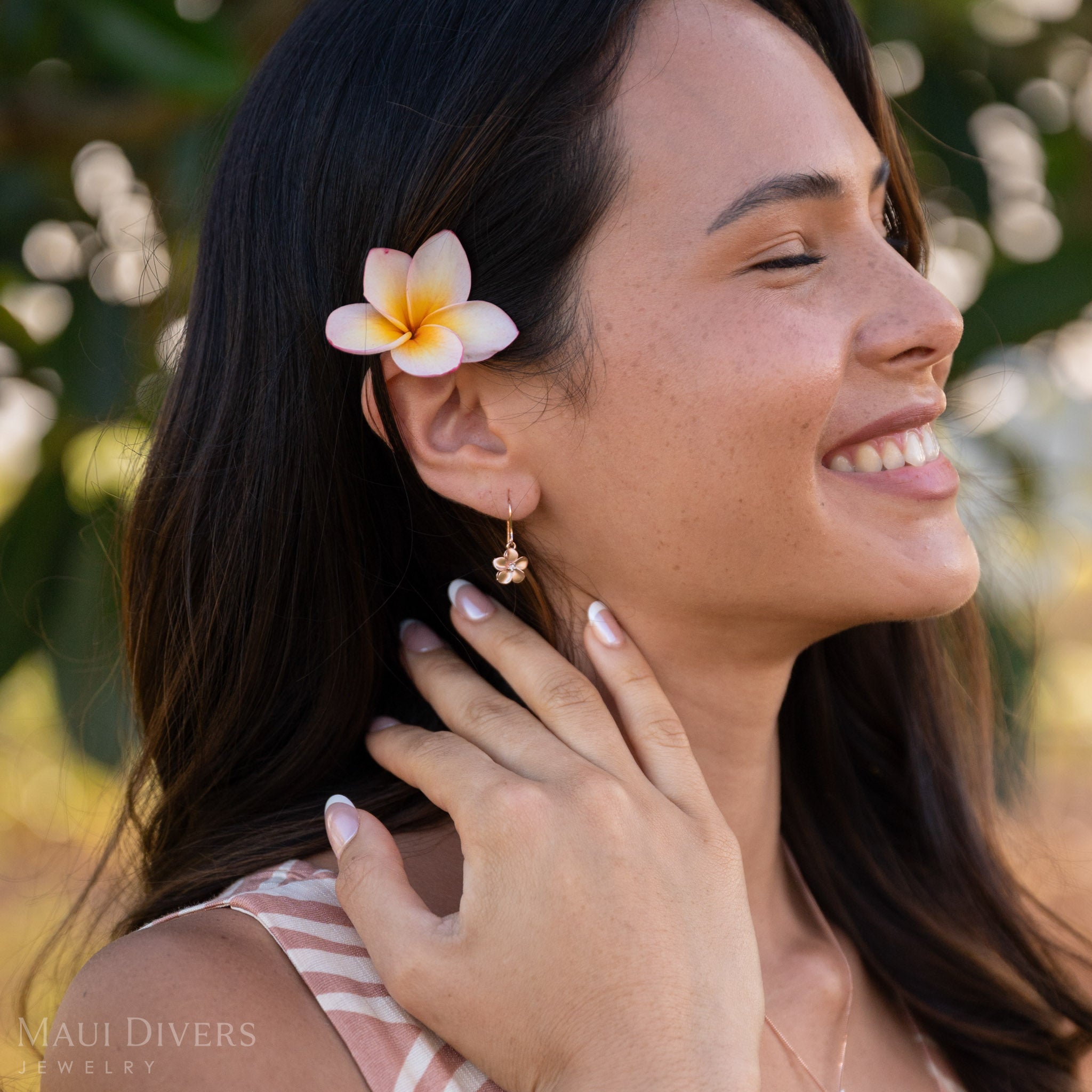 Close-up of a smiling woman with a plumeria flower in her hair wearing a Plumeria Earring with a diamond in 14k rose gold, against a blurred leafy green background