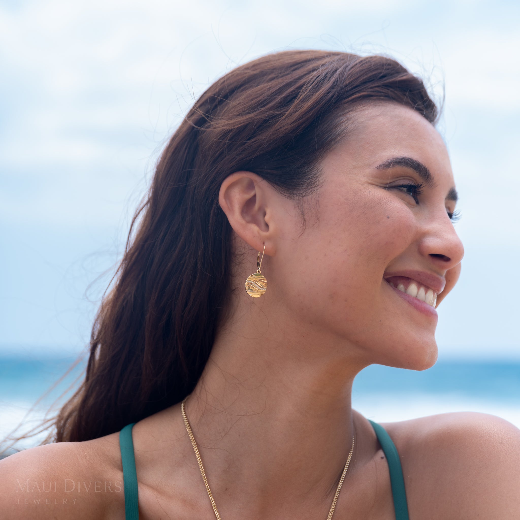 Woman wearing gold and diamond round ocean sand earrings and a gold chain with a blurred beach background