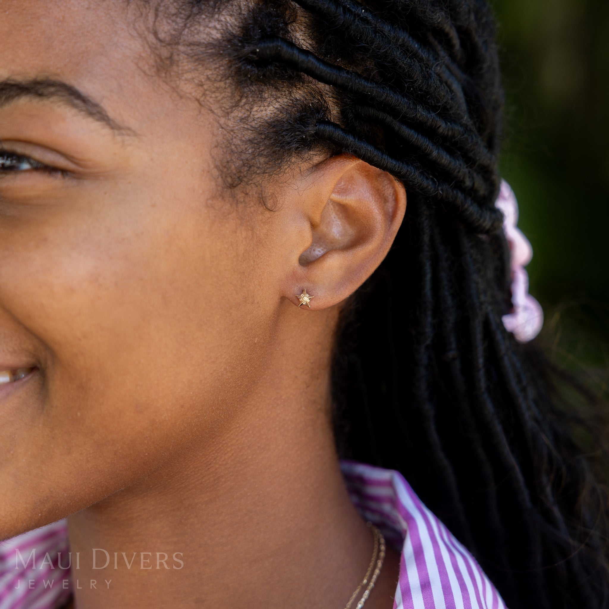 A side view of a smiling woman in a pink and white striped top wearing a Starfish Earring in 14k yellow gold, against a blurred green background