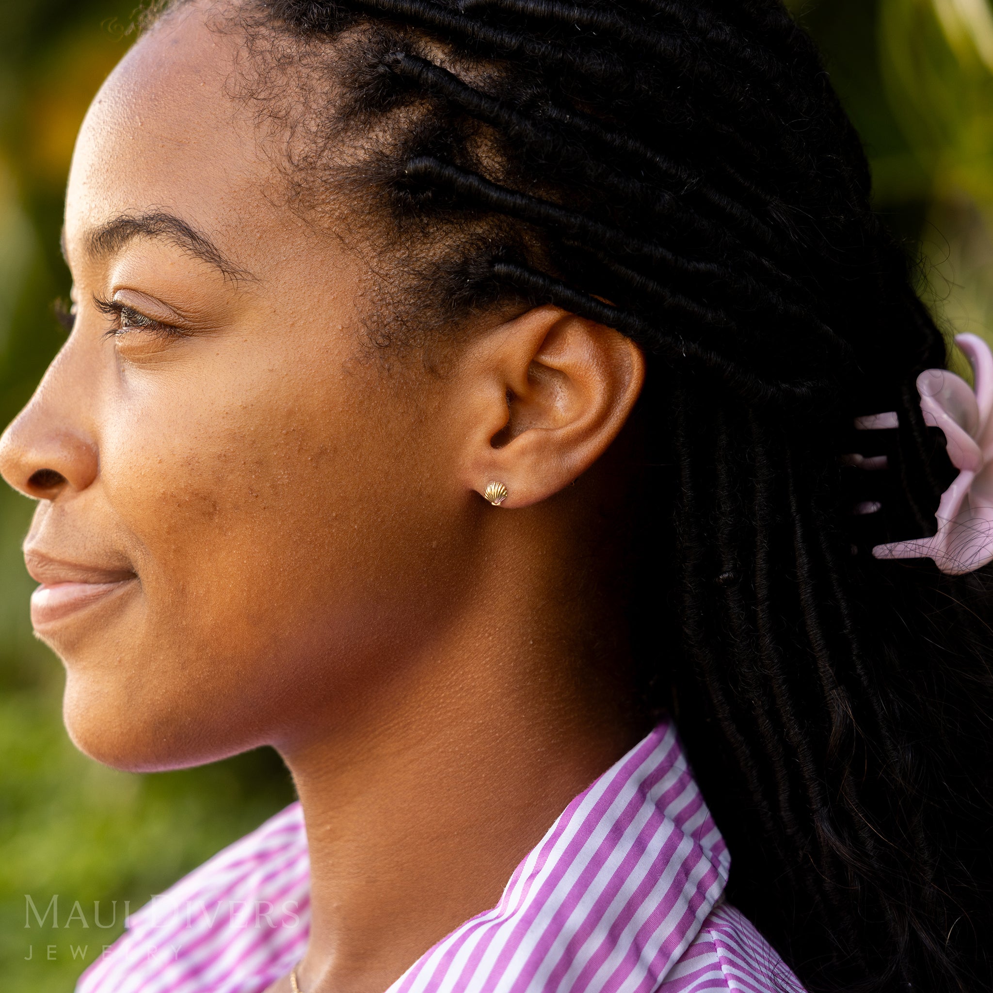 A side view of a smiling woman in a pink and white stripe top wearing a Hawaiian Sunrise Shell Earring in 14k yellow gold, against a blurred leafy green background