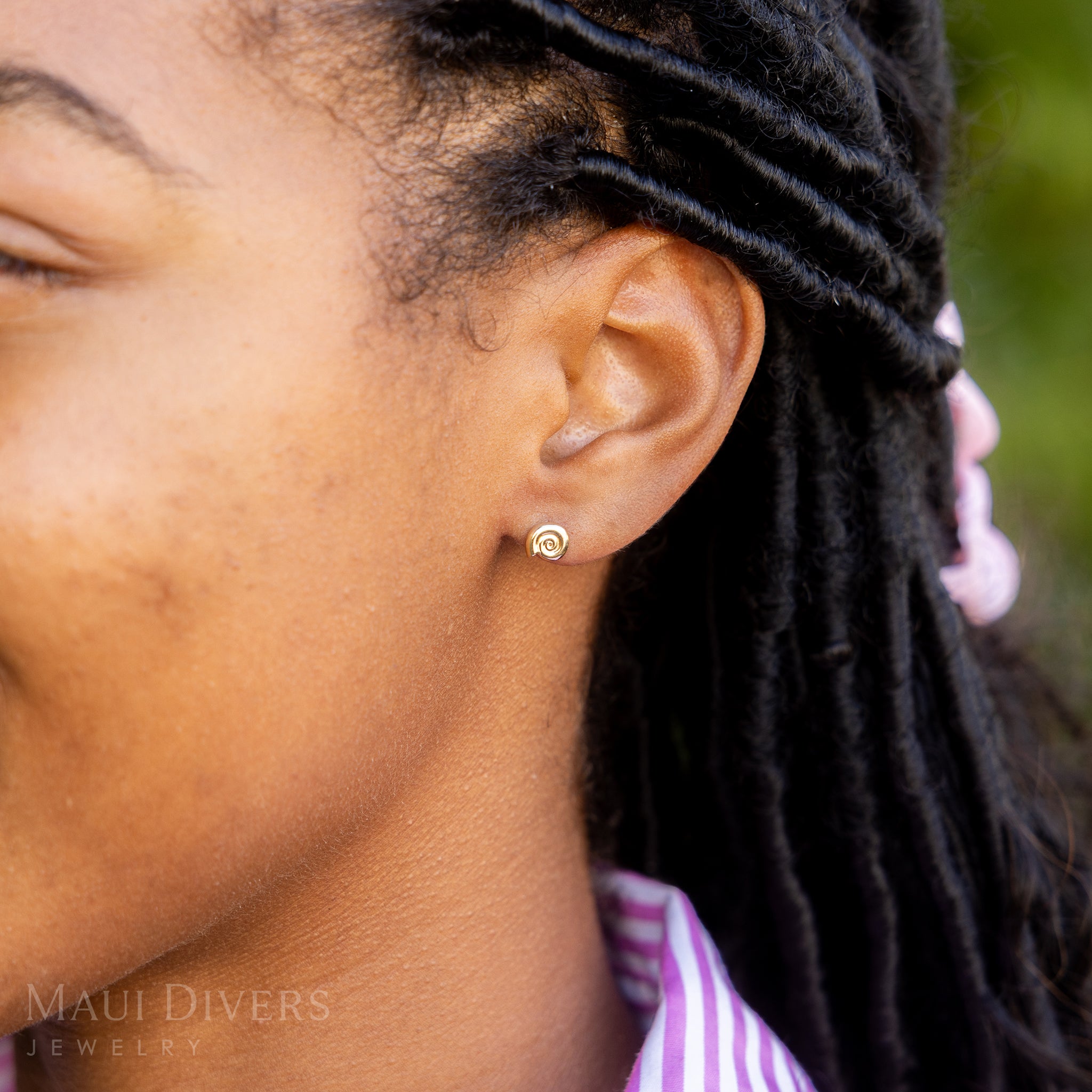 A side view close-up of a woman in a pink and white stripe top wearing a Conch Shell Earring in 14k yellow gold, against a blurred green background
