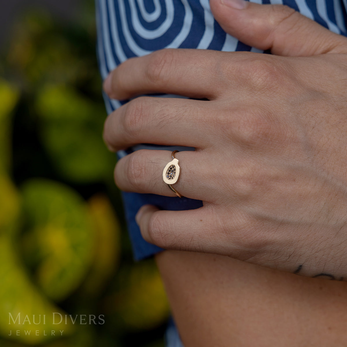 A close-up of a person wearing a Cute Fruits Papaya Pendant in Ring in 14k yellow gold with black diamonds on their hand, against a blurred green leaf background