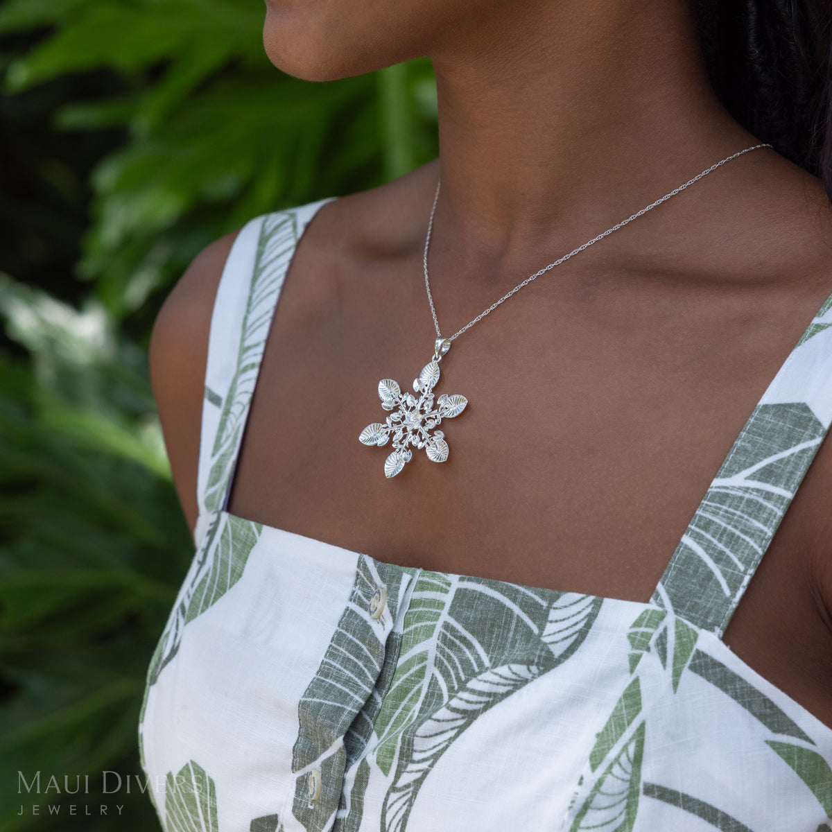 A close-up lifestyle shot of a model wearing a silver snowflake pendant necklace, styled with a green and white tropical leaf print top against a blurred green foliage background.