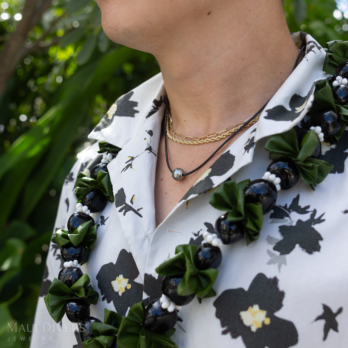 A close-up shot of a man wearing layered gold necklaces and a black coral tahitian black pearl necklace with a lei around him and a patterned white shirt against a blurry nature background