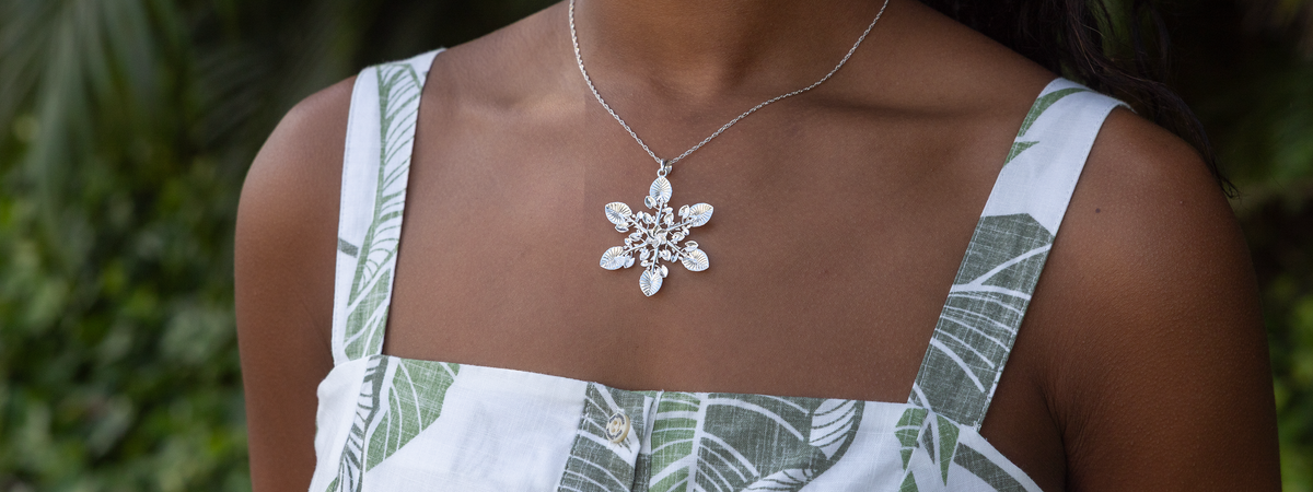 A sterling silver kalo leaf snowflake-style pendant necklace, worn by a model wearing a white and green tropical print top against a blurred green background.